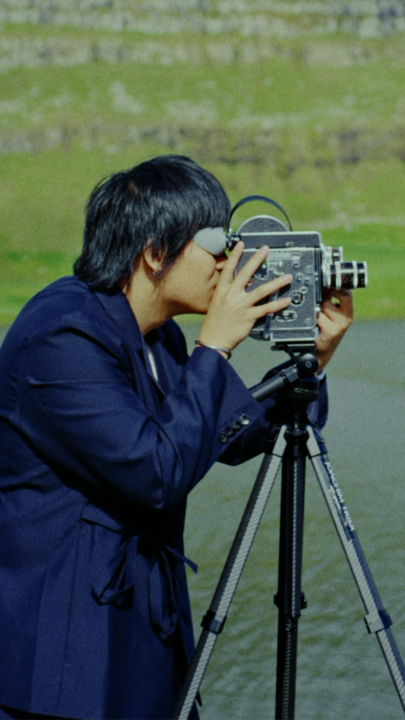 A young man with dark hair in a navy blue jacket looks through a vintage film camera mounted on a tripod, with a stone wall and grassy area visible in the background.