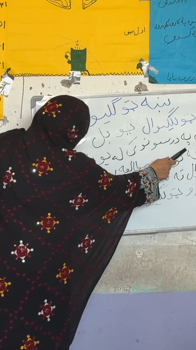 A woman in a black and red patterned hijab writes Arabic script on a whiteboard in what appears to be a classroom setting.