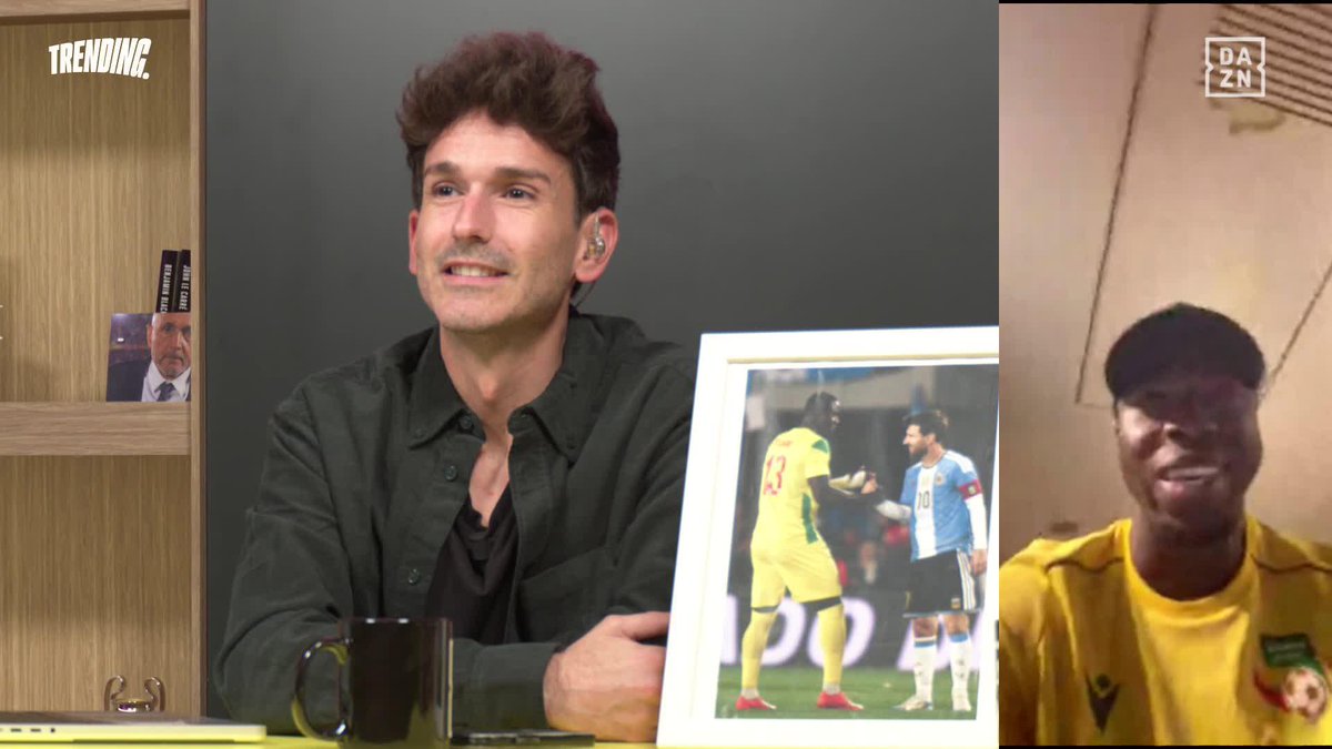 A bearded TV host in a black shirt sits smiling at a desk displaying a framed photo of soccer player Tijani (#13 in yellow) shaking hands with Lionel Messi (#10 in Argentina blue), while Tijani appears in a yellow jersey on a video call on the right. The image illustrates the "TRENDING" moment mentioned in the post, as the host showcases the historic photo that Tijani plans to display next to his bed.
