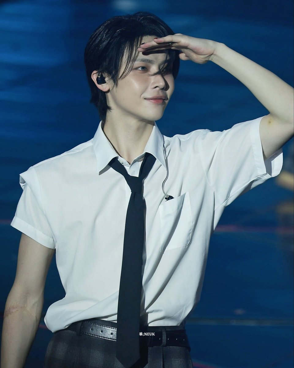A young man in a white short-sleeved shirt and black tie shields his eyes with his hand against a blue background, with the text '影:NEUK' at the bottom.