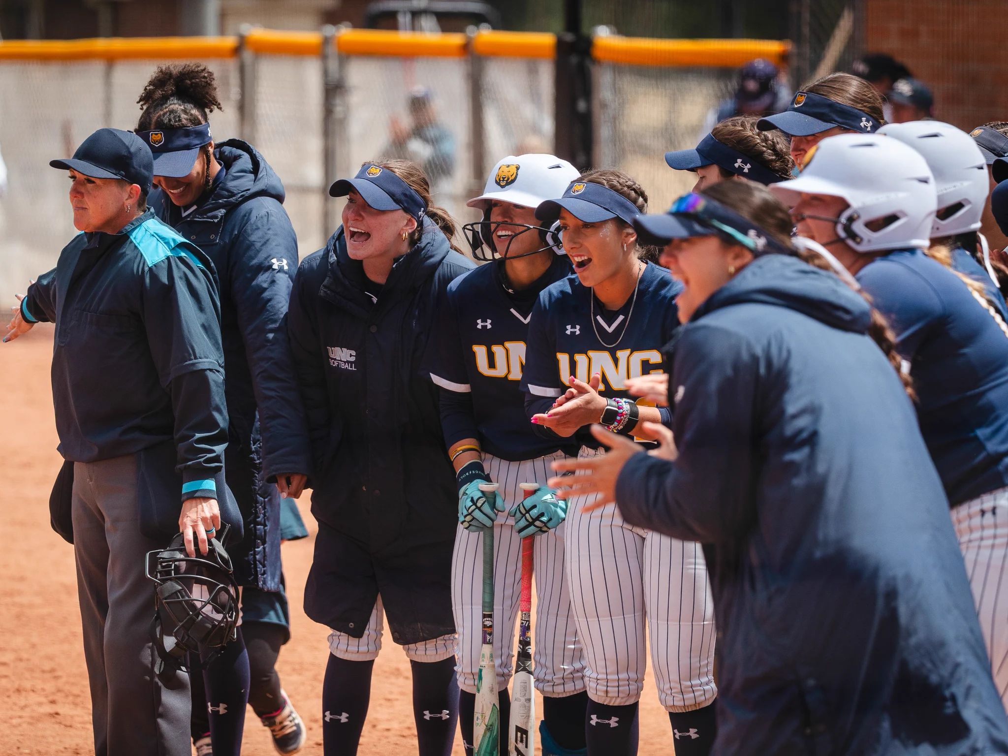 A group of UNC athletes stands on a softball field. They are wearing uniforms and baseball caps, some holding bats. Coaches and team members are gathered around, appearing enthusiastic and engaged. The setting is a sunny day at a softball game.