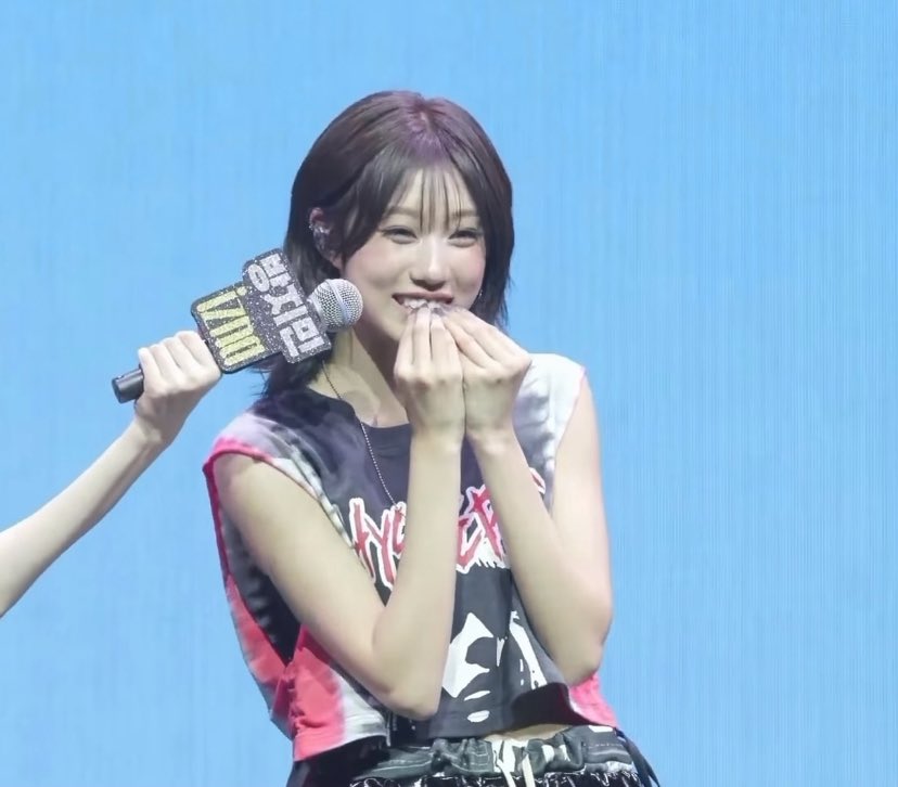 A young woman with short hair laughs while covering her mouth, holding a microphone labeled 'M COUNTDOWN' at a K-pop music show.