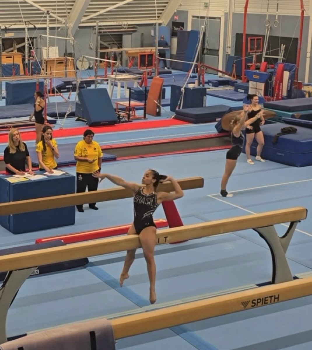 A gymnast wearing a "BRASIL" leotard sits on the "SPIETH" balance beam while performing, as coaches in yellow shirts observe and other athletes warm up nearby in the training facility. The setting appears to be a Brazilian gymnastics training session, consistent with the post featuring Thais Fidelis, a Brazilian gymnast.