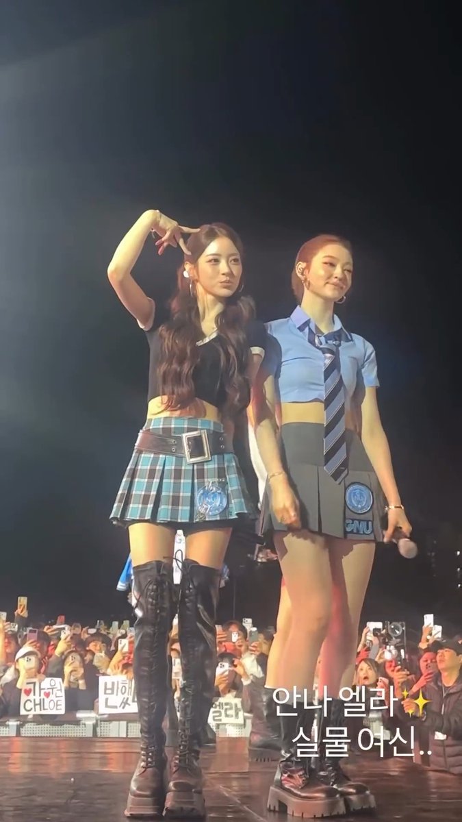 Two female performers pose on stage at a concert, one making a peace sign over her head while wearing a black crop top, plaid skirt, and knee-high boots, and the other in a blue crop top with a striped tie and gray pleated skirt. A fan sign reading "I ❤ CHLOE" and Korean text is visible, with overlaid Korean text "안나 엘라✨ 실물 역시.." (Anna Ella✨ she really is the real deal..) praising the two members' real-life appearance.