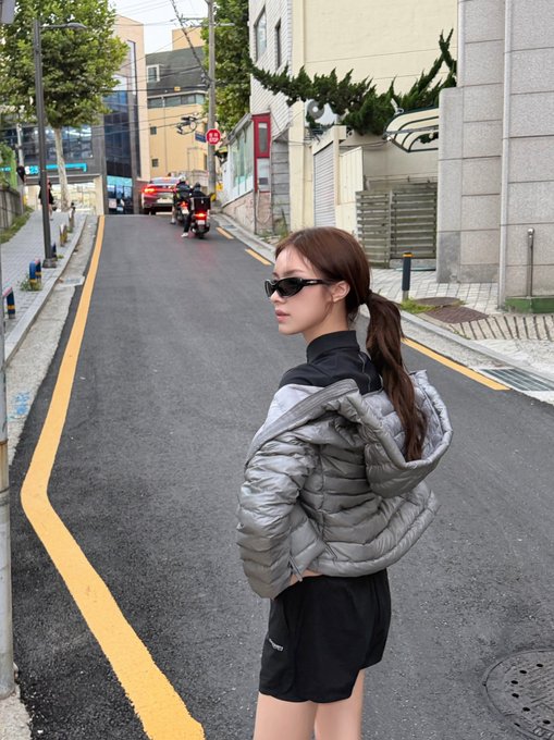 A young woman in sunglasses and a silver puffer jacket stands on a sloped street, looking back over her shoulder toward the camera.
