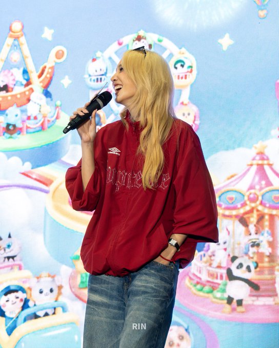 A blonde woman stands smiling while holding a microphone, wearing a maroon jacket with a logo and 'RIN' printed near the bottom, set against a colorful cartoon amusement park backdrop.