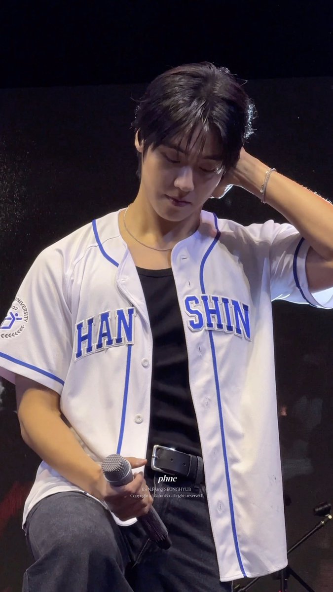 A young man in a white and blue 'HAN SHIN' baseball jersey over a black shirt holds a microphone, with his hand touching his hair on a dark stage.