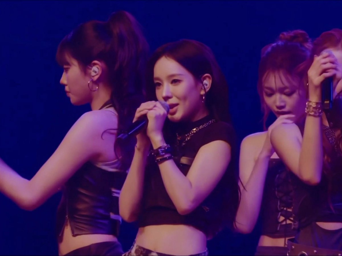 A female performer sings into a microphone on stage, flanked by two backup dancers in black outfits, all illuminated by deep blue stage lighting.