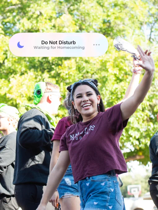A person is smiling and waving during an outdoor event, wearing a maroon t-shirt with "NM State Aggies" printed on it. Other people are nearby, some wearing green accessories. A notification at the top reads "Do Not Disturb: Waiting for Homecoming." Lush green trees are in the background.