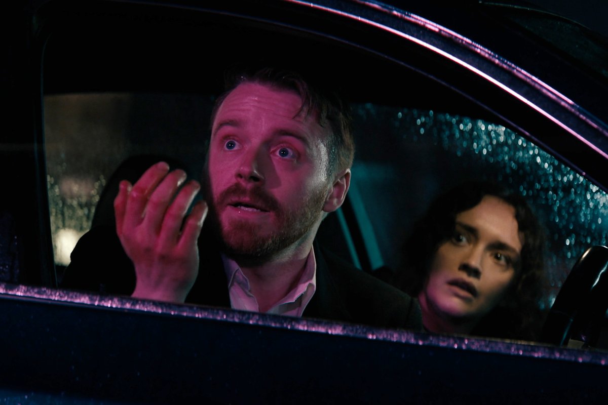 Two people sit inside a car at night, illuminated by pink and blue neon light, with the bearded man in the foreground looking startled while pressing his hand against the rain-speckled window—capturing a tense, rain-soaked moment befitting the espionage thriller "Slow Horses."