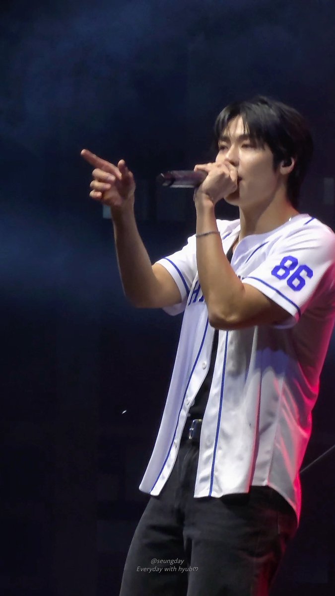 A performer in a white baseball jersey with the number 86 sings into a microphone on a dimly lit stage, pointing outward with one hand.