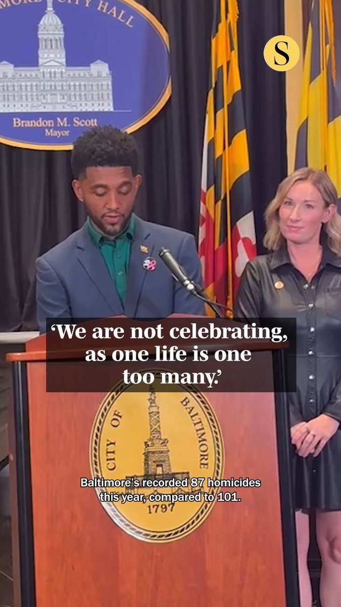 Baltimore Mayor Brandon M. Scott stands at a podium with the City of Baltimore seal, flanked by a Maryland flag, while text reads 'We are not celebrating, as one life is one too many.' and 'Baltimore's recorded 87 homicides this year, compared to 101.'