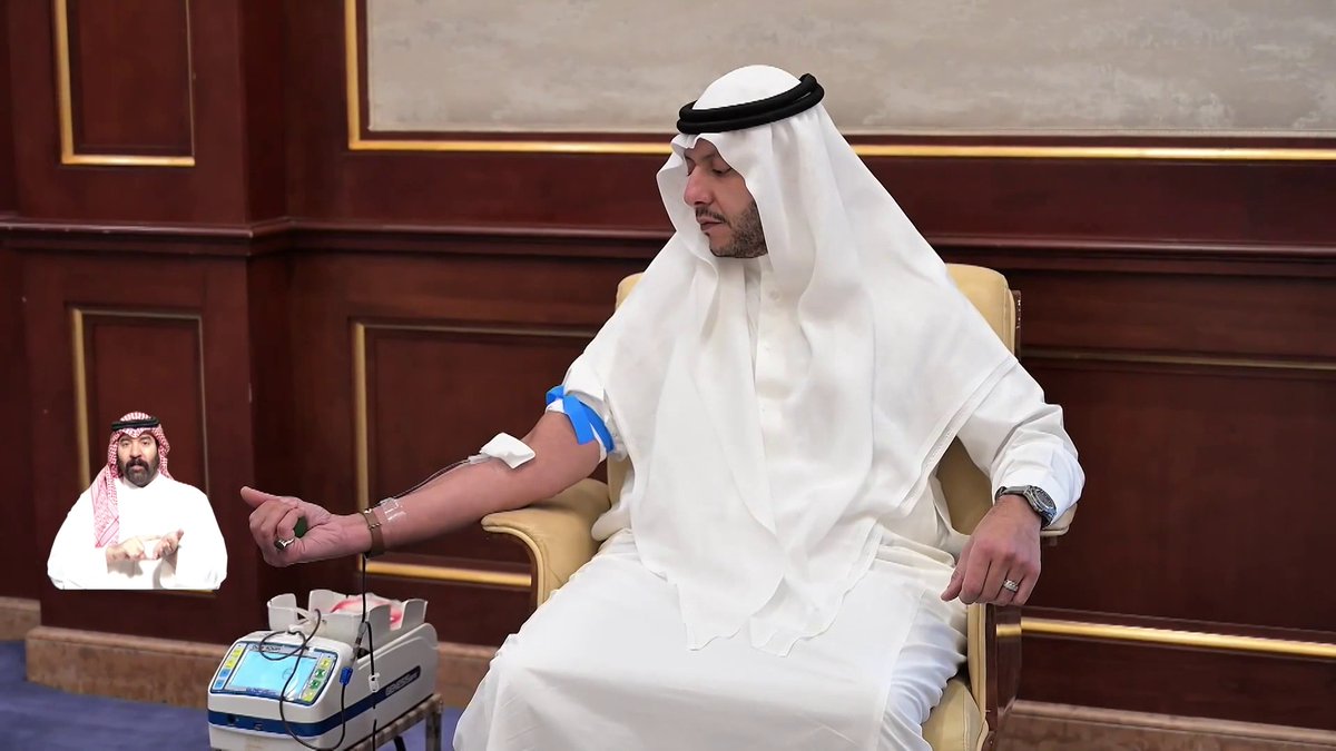 A man in traditional white Middle Eastern attire (thobe and ghutra) seated in a chair while having blood drawn into a medical device, with a sign language interpreter in the bottom left corner.