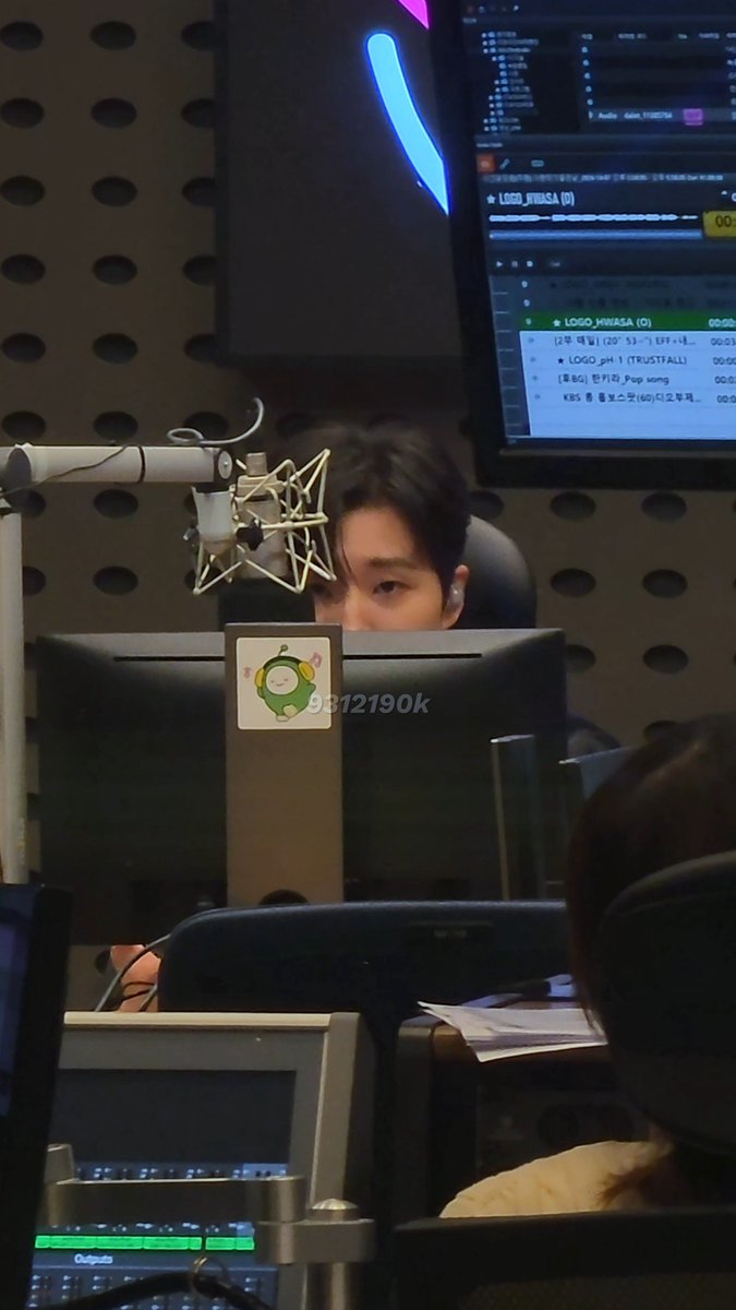 A person is seen behind a microphone and computer monitors in what appears to be a radio studio, with a monitor displaying 'LOGO_HWASA' track listings.