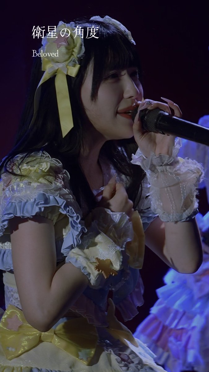 A young woman in an ornate yellow and white frilly dress singing into a microphone, with overlaid text "衛星的角度" (Satellite Angle) and "Beloved."