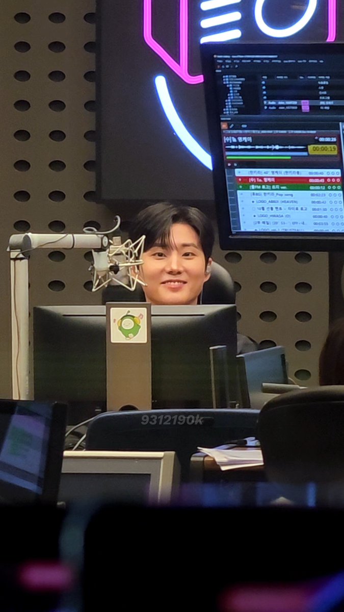 A man in a radio broadcasting studio smiles behind a microphone, with computer monitors displaying audio editing software visible above him.