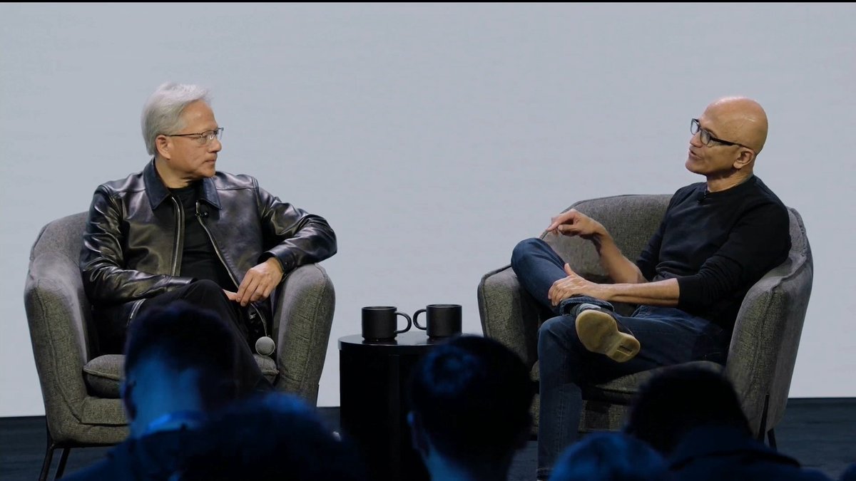 NVIDIA CEO Jensen Huang and Microsoft CEO Satya Nadella sit in armchairs on stage, engaged in a fireside chat discussion. Two mugs rest on a small table between them, with audience members visible in the foreground, fitting the context of their conversation reflecting on their early collaboration building supercomputers.