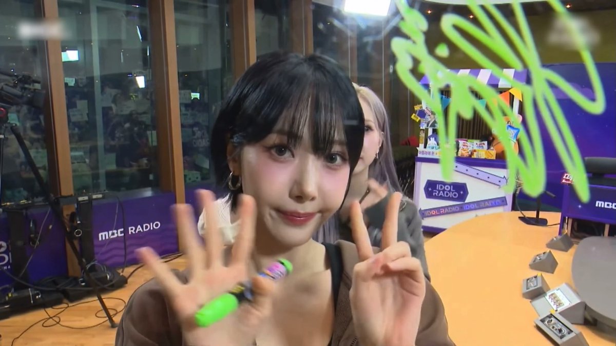 A woman with dark hair and bangs makes a peace sign and gestures toward the camera in what appears to be a radio studio, with 'MBC RADIO' and 'IDOL RADIO' visible in the background.