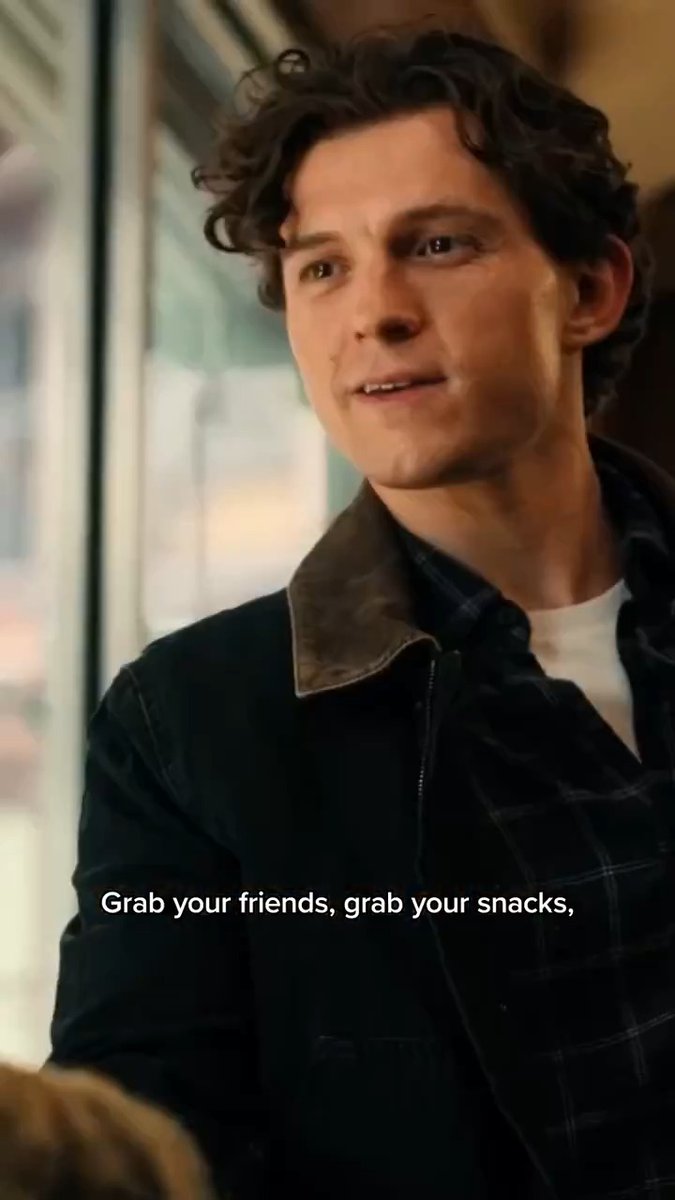 A young man with curly dark hair smiles in a warmly lit indoor setting, wearing a dark jacket over a plaid shirt. The caption "Grab your friends, grab your snacks," is displayed at the bottom, promoting the Spider-Man: Brand New Day movie release.