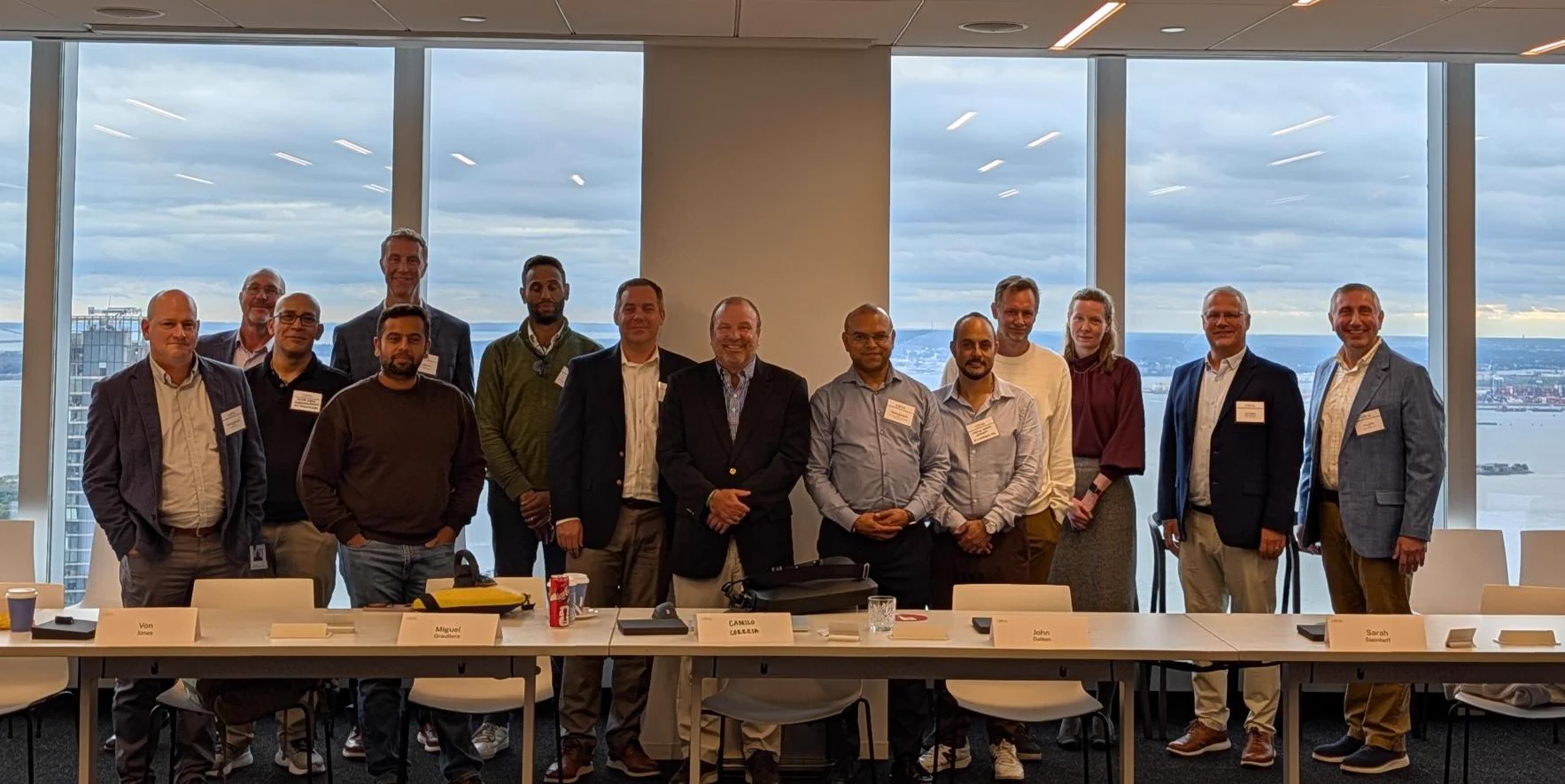 A group of people standing in a conference room with large windows showing a cityscape in the background at the Citrix FSI roundtable. There are name tags on some individuals, and a long table with chairs in the foreground.