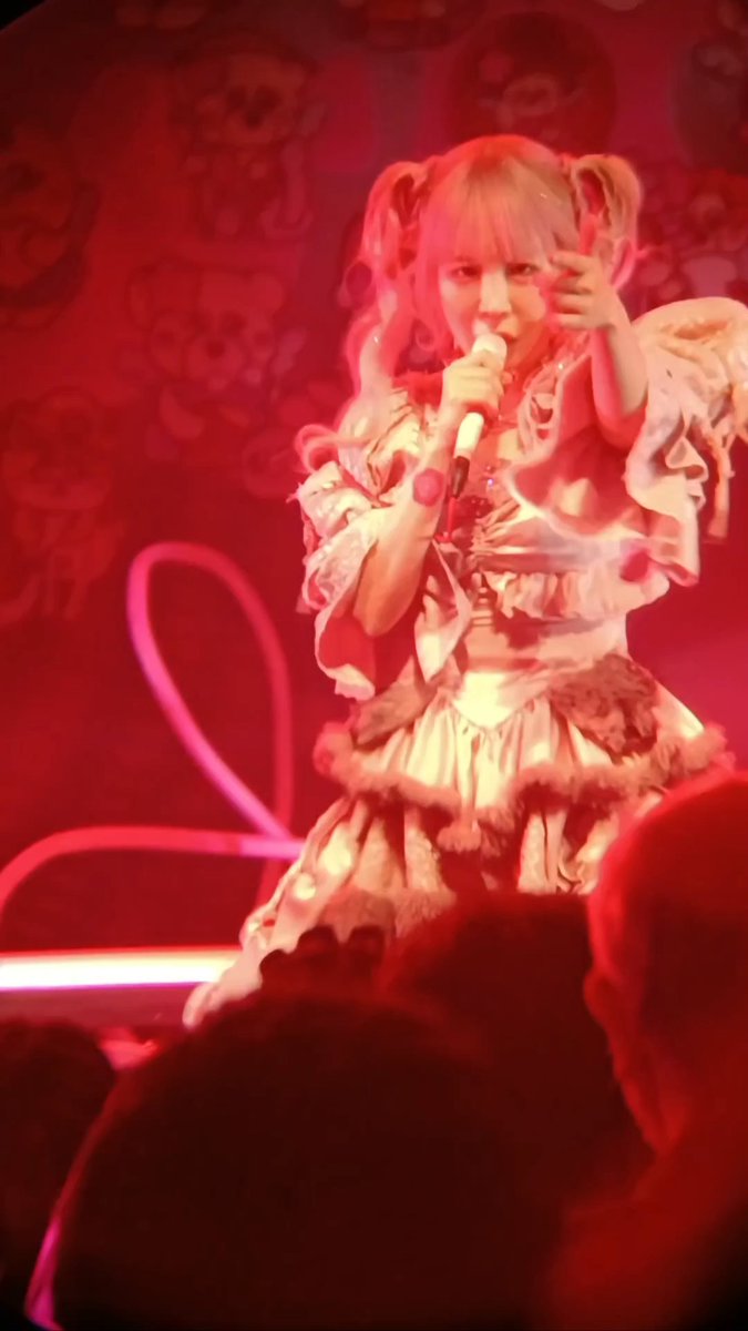 A young female performer with pink pigtails on stage in an elaborate frilly outfit, holding a microphone under dramatic red lighting with audience members visible in the foreground.