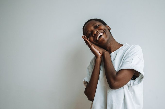 A photo of a young Black woman in a white t-shirt against a plain light gray background, smiling brightly with her hands cupped against her cheek in a joyful pose.