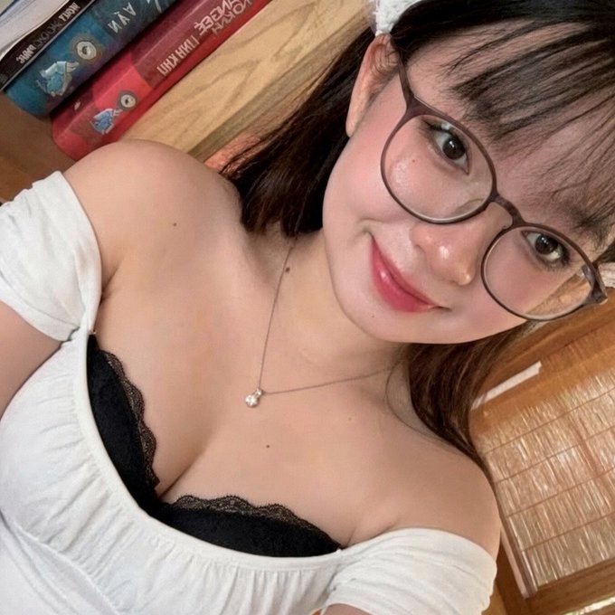 A selfie of a young woman wearing round glasses, a white off-shoulder top with a black lace bra underneath, and a delicate necklace, posing in front of a bookshelf with Pokemon-related items.