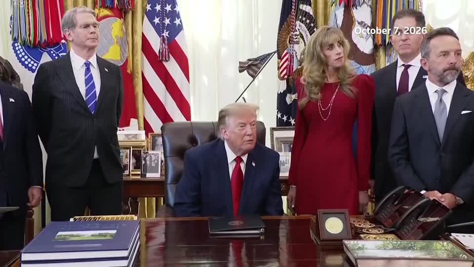 A group of officials, including a woman in a red dress, stand behind a seated President Donald Trump at the Resolute Desk in the Oval Office, with the date "October 7, 2026" displayed in the upper right corner. The image appears to capture the moment Trump made his remarks about Federal Reserve board members, with Chairman Kevin Warsh likely among those present.
