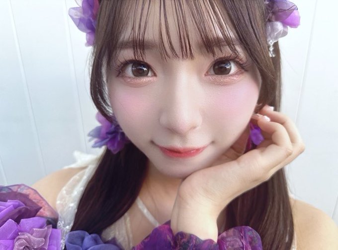 A young woman with brown hair and heavy blush poses in a selfie, wearing purple floral accessories and a purple ruffled top.