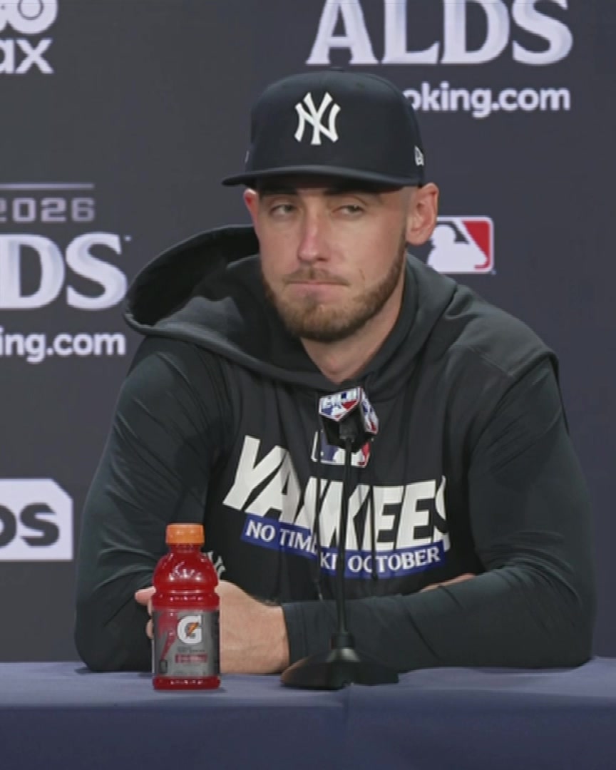A New York Yankees player, appearing to be Cody Bellinger, sits at a press conference table during the ALDS, wearing a Yankees cap and a black Yankees "NO TIME FOR OCTOBER" hoodie, with a Gatorade bottle in front of him. The image captures the post-game media setting referenced in the quote, where Bellinger praises Yankees manager Aaron Boone for upholding the franchise's storied legacy.