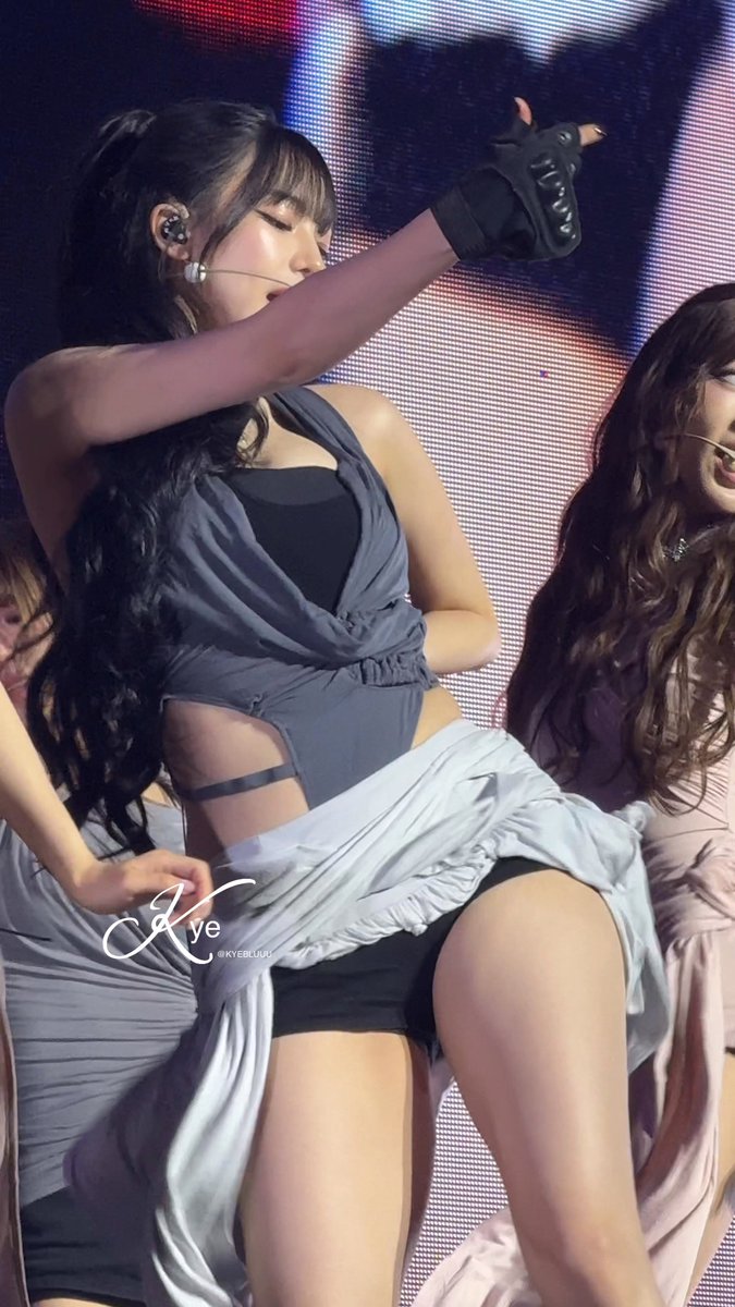 A concert photo of a female idol with long dark hair in a high ponytail, wearing a gray draped top, black shorts, and a glove with studs, performing on stage with other dancers partially visible.