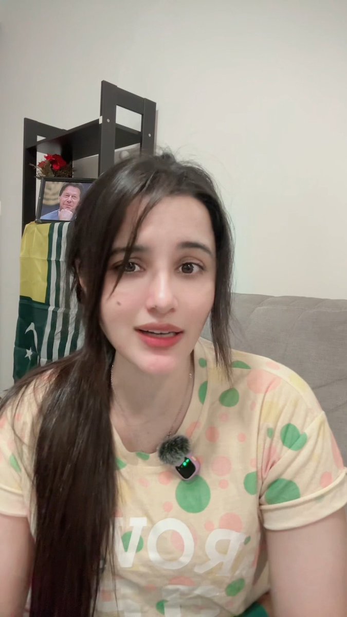 A young woman with long dark hair wearing a polka-dot shirt speaks to the camera, with a Pakistani flag and a photo of Imran Khan visible on a shelf behind her.