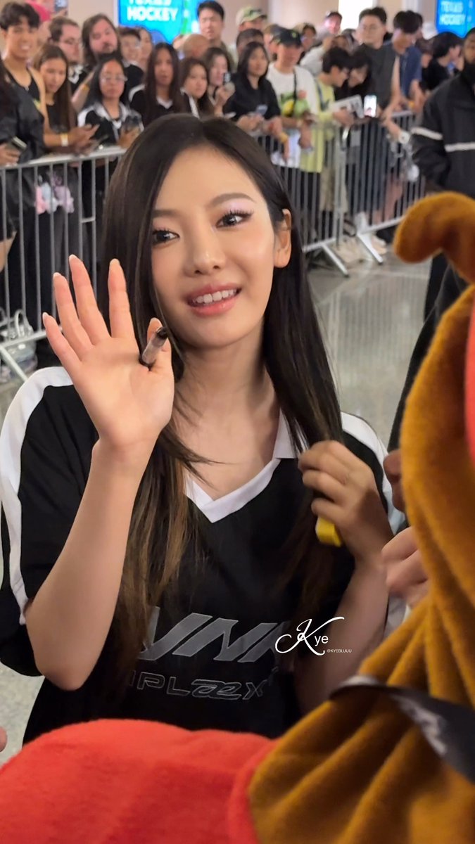 A young Asian woman (Yeji of ITZY) waves while holding a marker, wearing a black and white Nike sports jersey at what appears to be a fan meet-and-greet event.