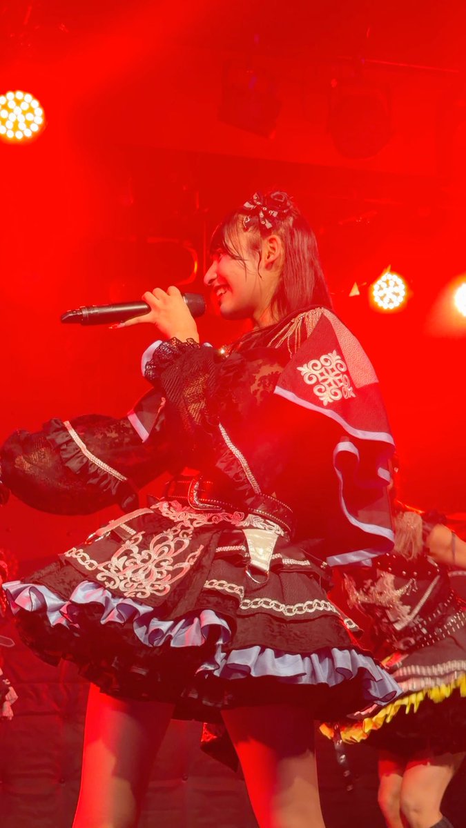 A young woman performs on stage bathed in red lighting, wearing an elaborate black gothic-lolita outfit and holding a microphone.