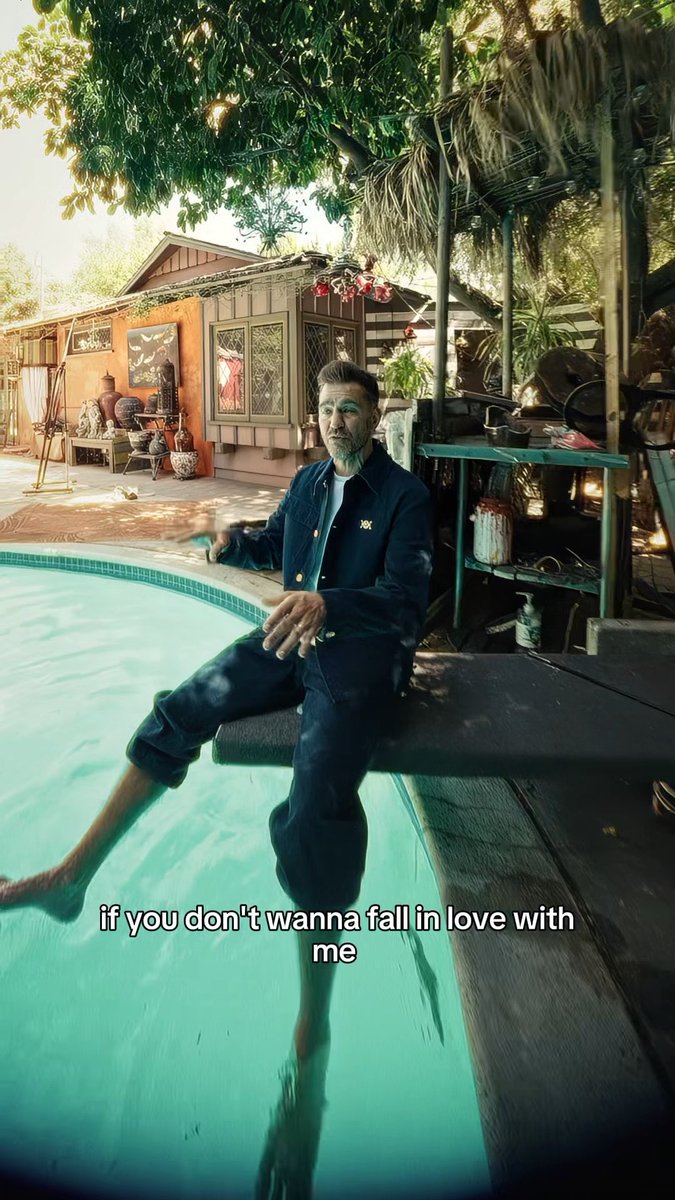 A man wearing a denim outfit sits on the edge of a turquoise swimming pool with his feet dipped in the water, with the text "if you don't wanna fall in love with me" displayed on screen.