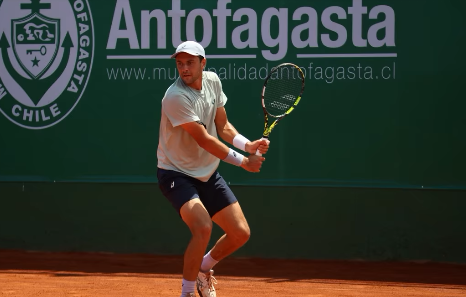 A tennis player wearing a white shirt, navy shorts, and white cap swings his racket on a clay court, with 'Antofagasta' and 'www.municipalidadantofagasta.cl' signage on the green wall behind him.