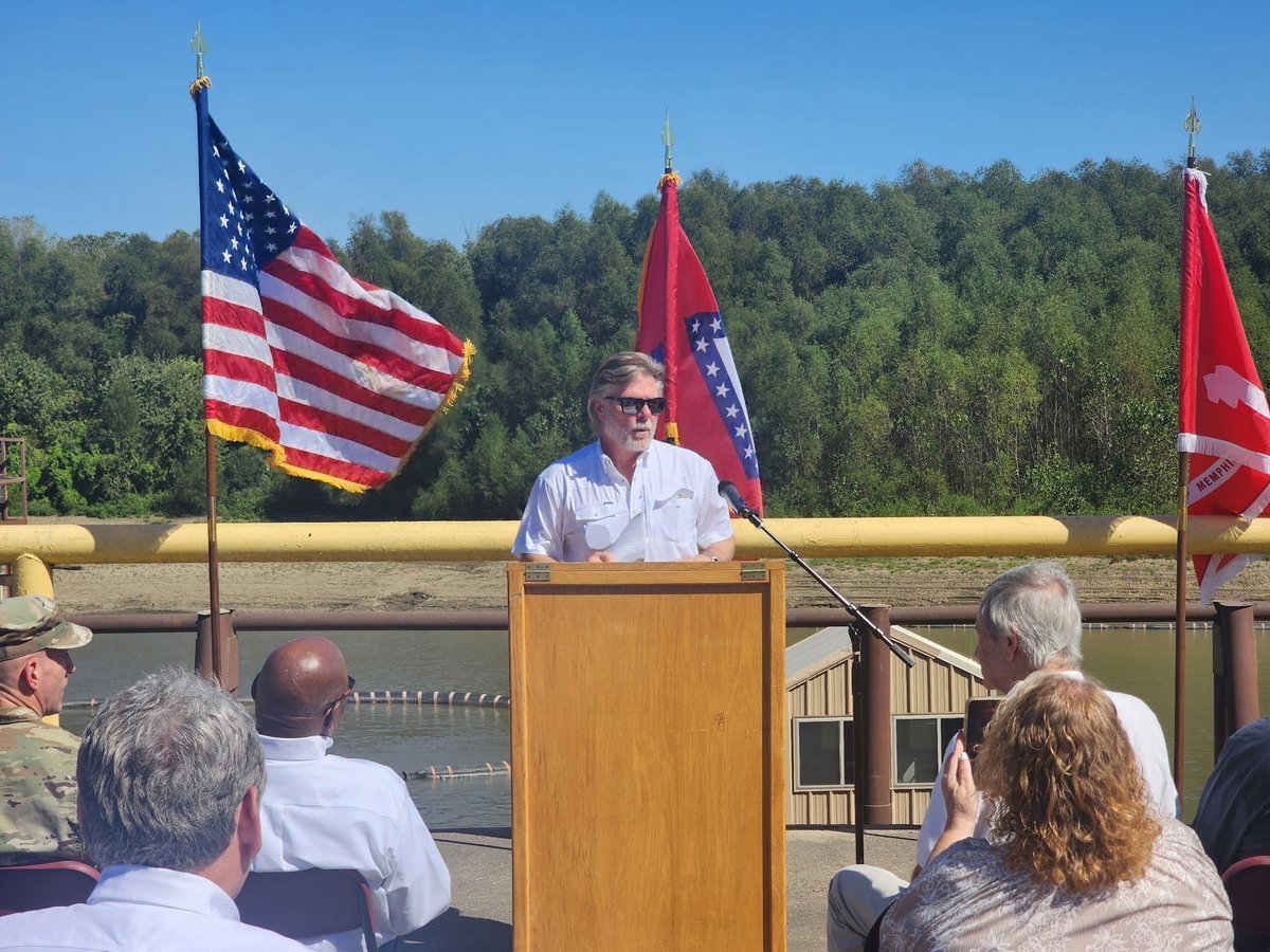 An outdoor ceremony is underway. A man wearing sunglasses and a white shirt speaks at a wooden podium with a microphone while an audience watches. An American flag and two red flags stand behind him, with trees and water in the background. Some flags have text.