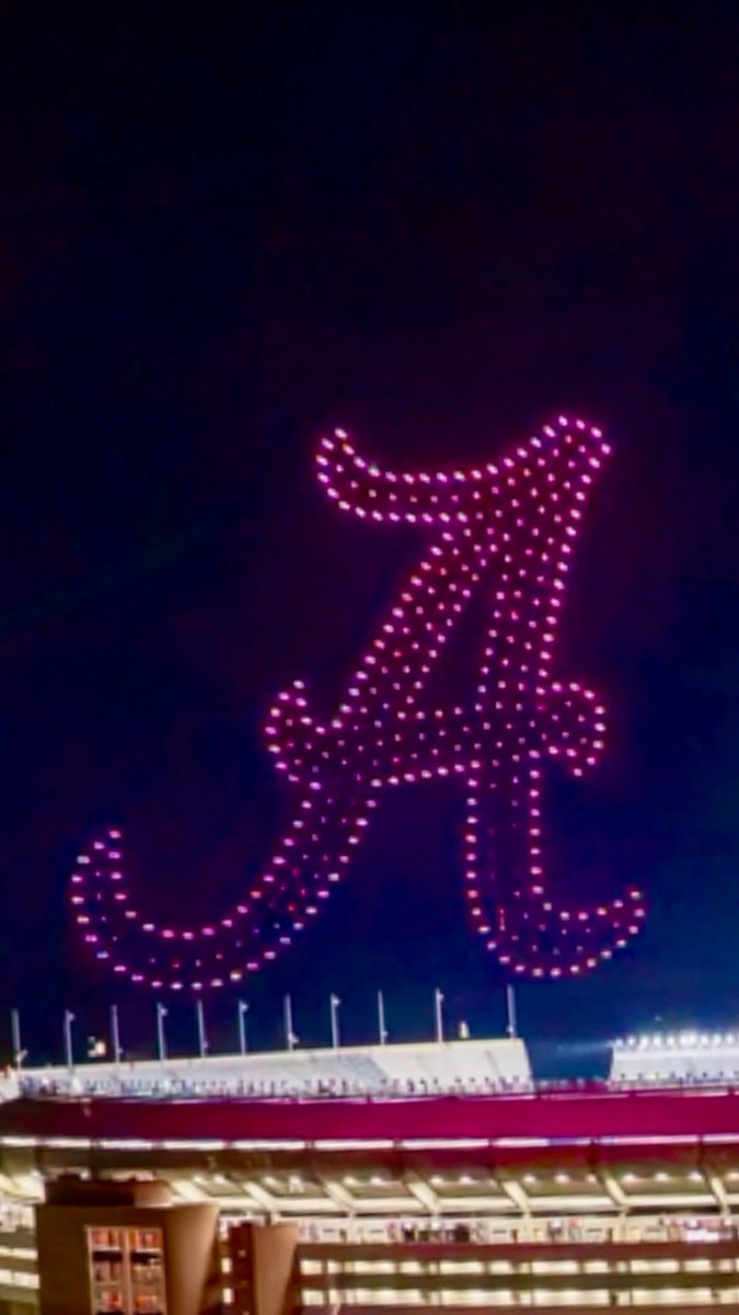 A nighttime view of a stadium with a drone light show forming the shape of a pink/purple cat or character in the sky above the venue.