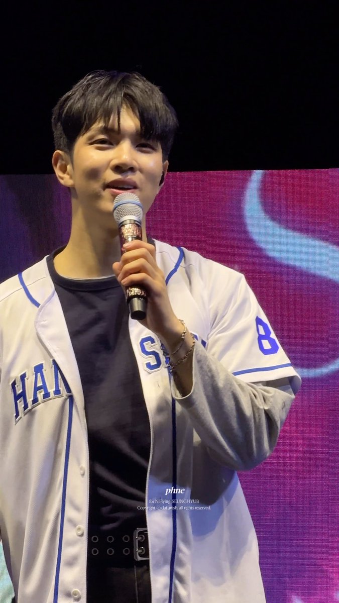 A young man with dark hair speaks into a microphone while wearing a white baseball jersey with 'HAN' and the number 8, over a black shirt.