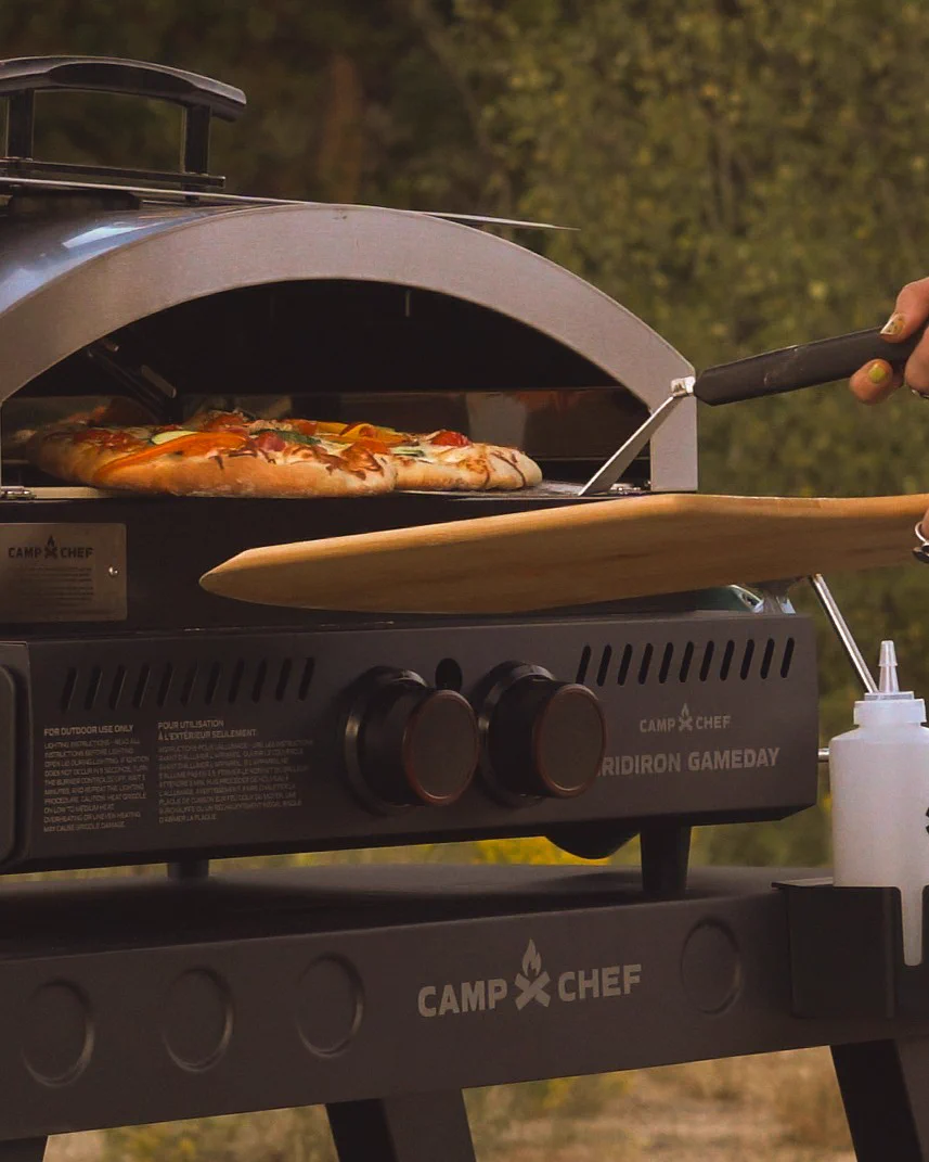 A person is using a Camp Chef outdoor pizza oven to bake a pizza. The scene is set outdoors with a blurred background of greenery. The pizza is being placed or retrieved using a wooden paddle.