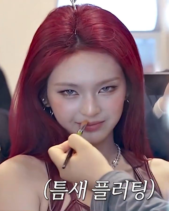 A close-up of a woman with bright red hair getting makeup applied to her lips, with Korean text "(틈새 플러팅)" overlaid at the bottom.