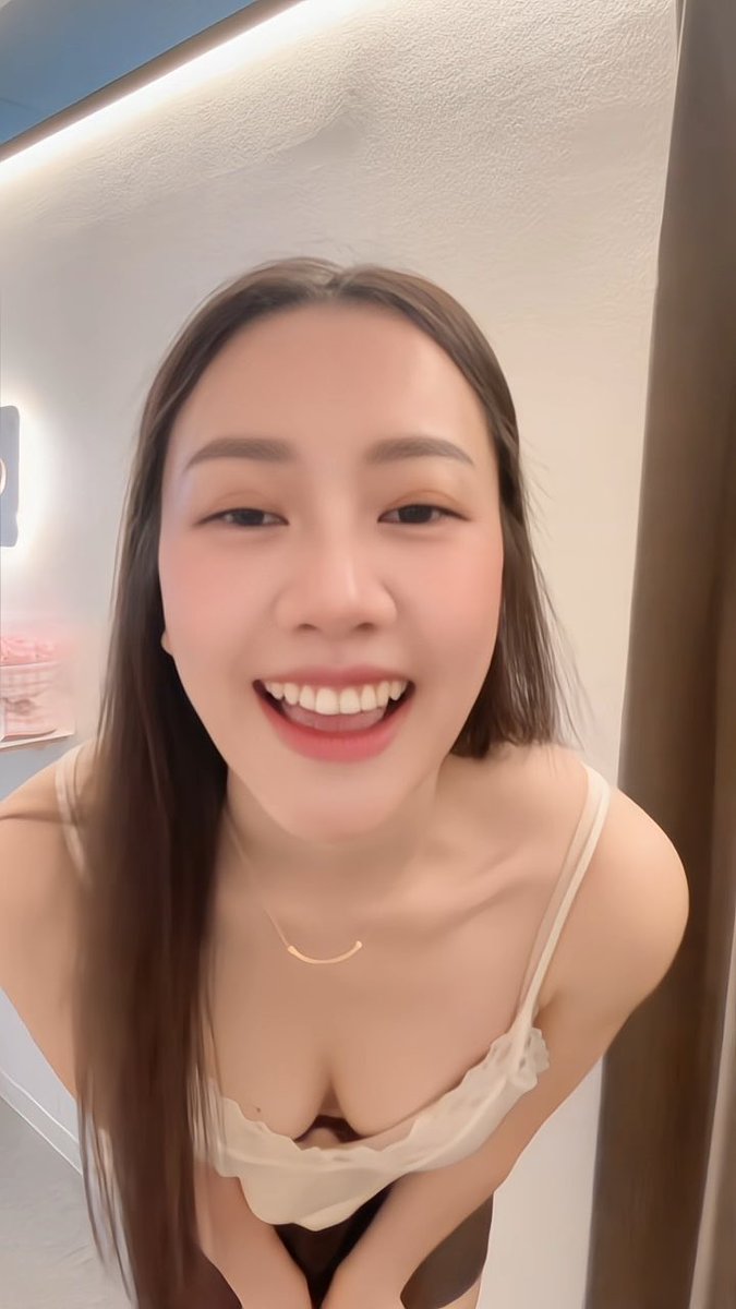A selfie-style video still showing a smiling young woman with long brown hair wearing a beige camisole top and a delicate gold necklace.