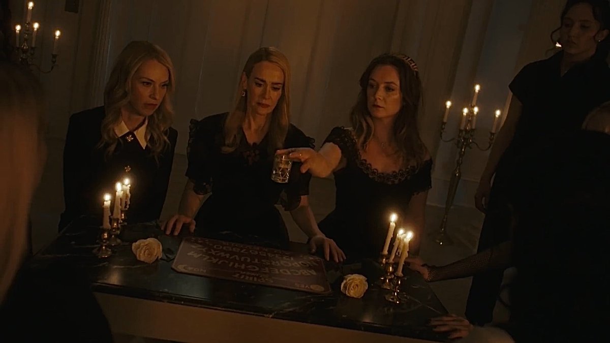 Three women in black dresses gathered around a lit altar with candles during what appears to be a ritual scene.