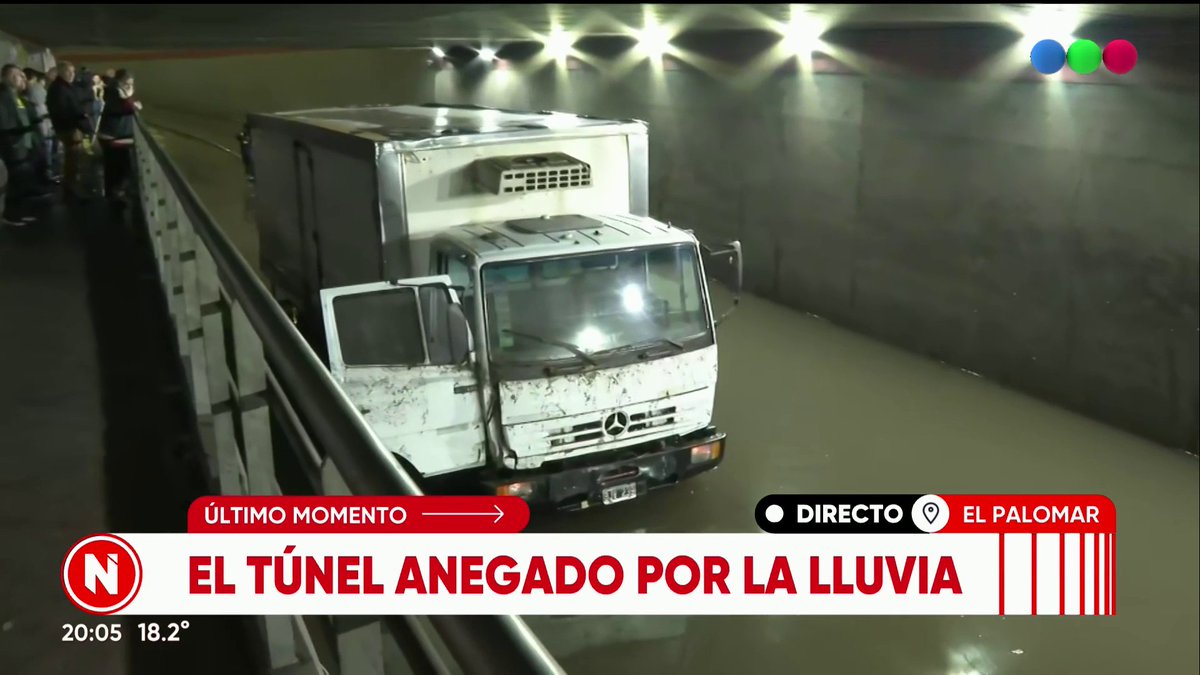 A news broadcast from Telefe (timestamp 20:05, 18.2°) shows a white Mercedes-Benz truck stranded in the flooded El Palomar tunnel, with onlookers watching from the walkway. The chyron reads "ÚLTIMO MOMENTO - EL TÚNEL ANEGADO POR LA LLUVIA" with a "DIRECTO EL PALOMAR" live indicator, illustrating the severe flooding mentioned in the post.