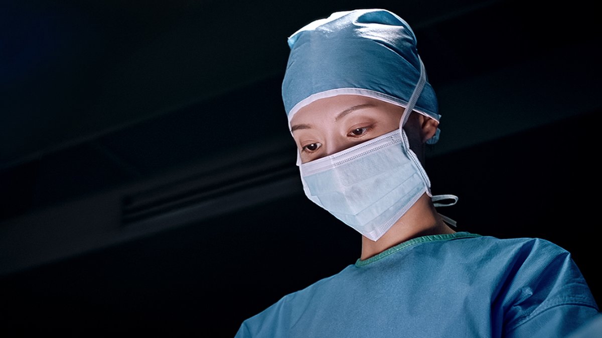 A close-up portrait of a female medical professional wearing a blue surgical cap, mask, and scrubs, looking toward the camera against a dark background.
