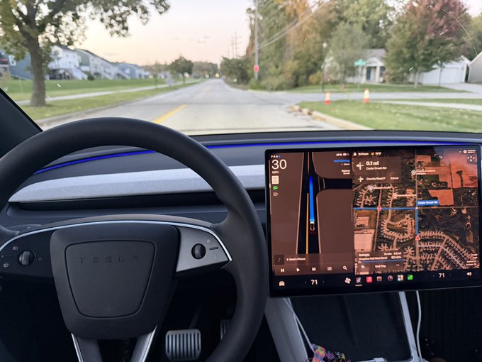 Interior view of a Tesla showing the steering wheel and center touchscreen displaying a navigation map, speed of 30 mph, and 71°F, with a residential street and orange traffic cones visible through the windshield.