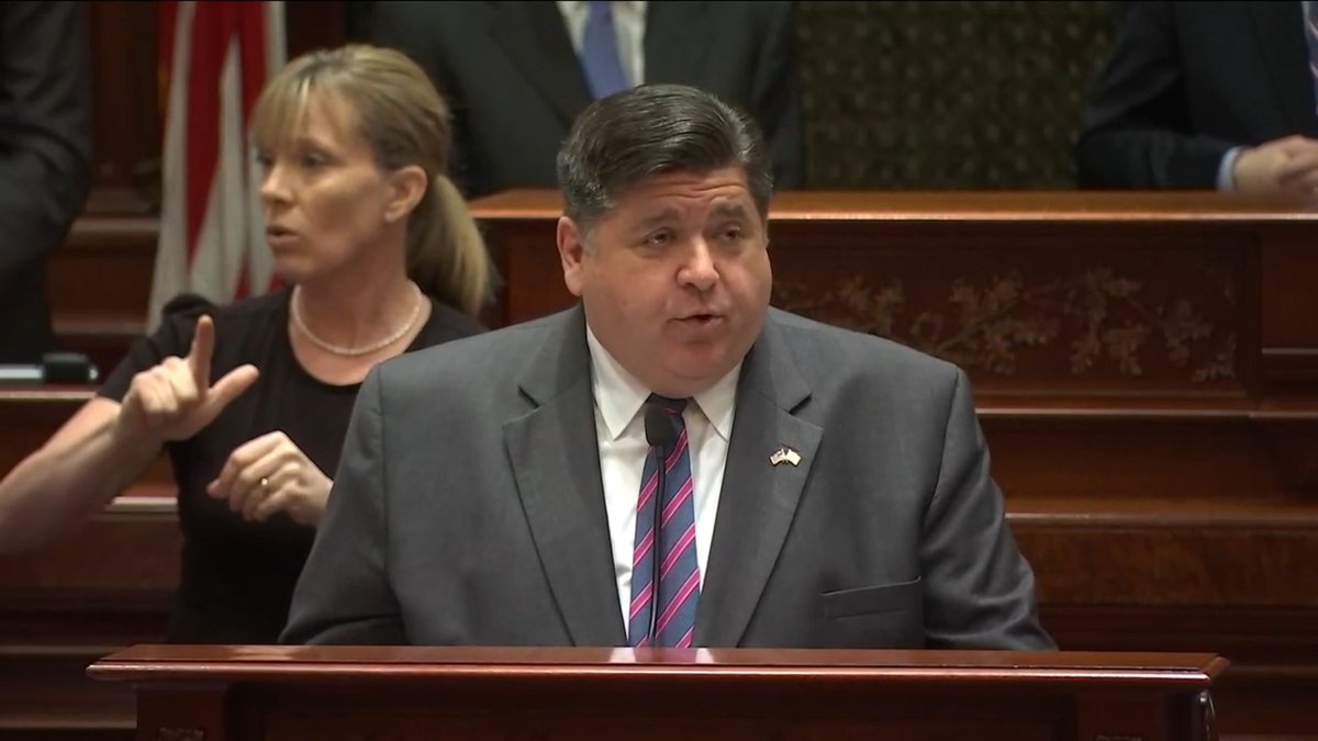 A man in a gray suit and striped tie stands at a podium speaking, while a woman in a black top and pearl necklace stands behind him making a hand gesture, in what appears to be a legislative chamber.