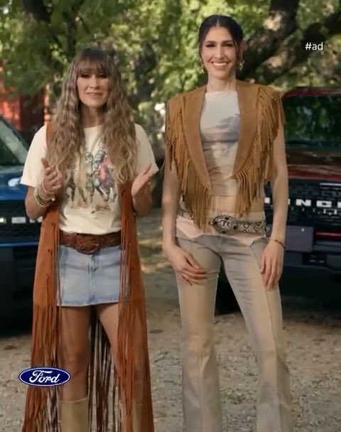 Two women wearing western-style clothing with fringe vests and belts pose next to a Ford Bronco, with a Ford logo visible and '#ad' in the corner.