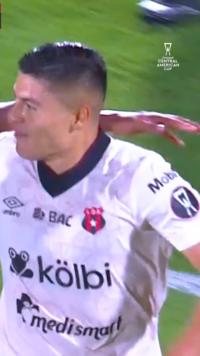 A soccer player wearing a white LD Alajuelense jersey featuring sponsor logos including "Kölbi," "BAC," "Medismart," "Umbro," and "Mobil," celebrates on the pitch during a Concacaf Central American Cup match, likely after scoring one of the team's goals on their journey to the semifinals.