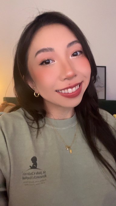 A selfie of a smiling young Asian woman with long dark hair, wearing a green sweatshirt and gold necklace with hoop earrings.